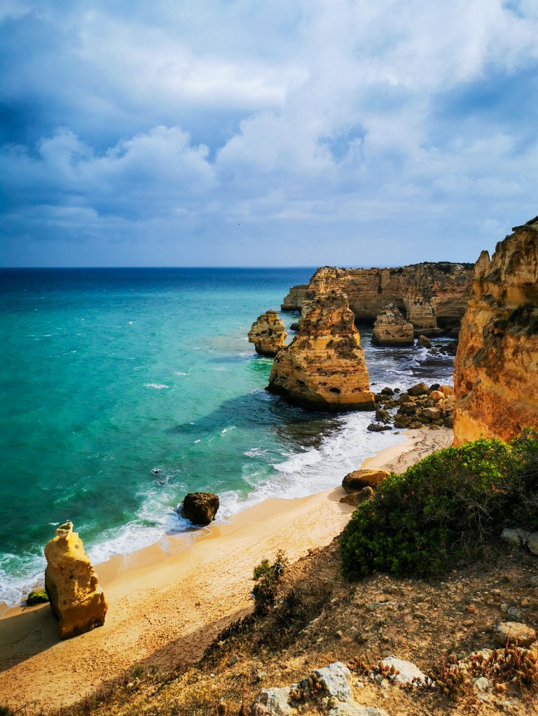 Praia da Marinha w pochmurny dzień z klifami, piaskiem i wzburzonym turkusowym oceanem.