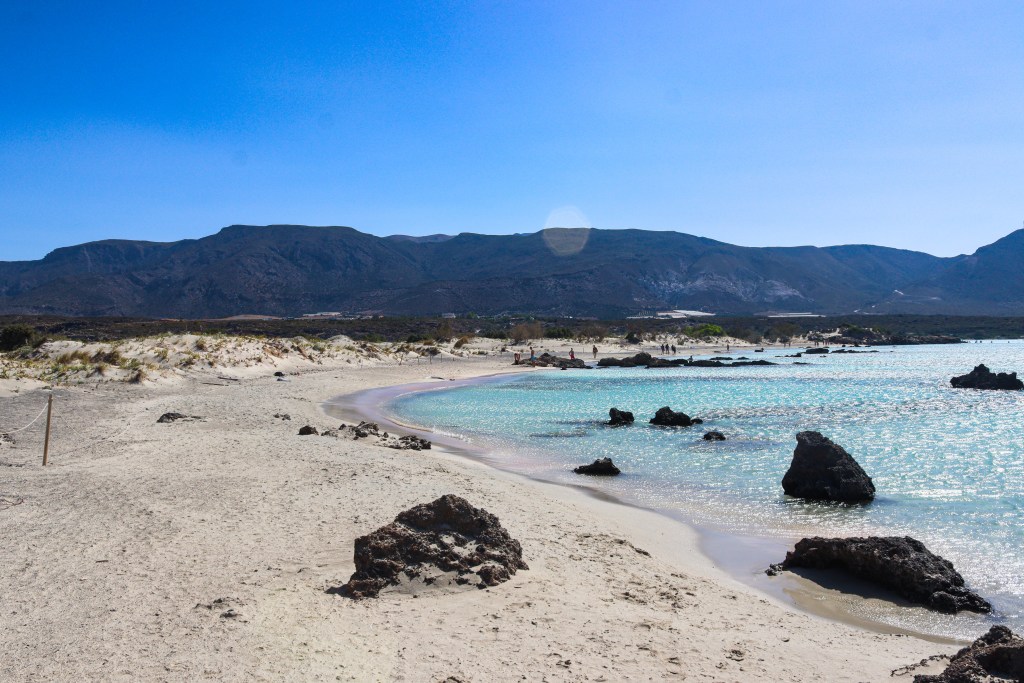 Elafonisi – Your Travel Guide to Crete’s Famous Pink Beach View of Elafonisi Beach with stones along the shore, turquoise waters and mountains in the background