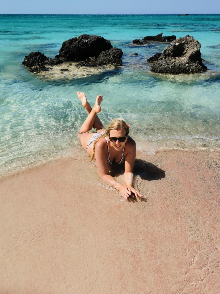 Elafonisi – Your Travel Guide to Crete’s Famous Pink Beach Woman lying on the pink sands of Elafonisi Beach, surrounded by turquoise waters and dark rocks