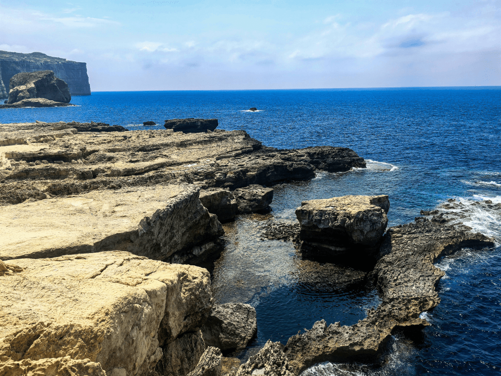 Exploring Gozo in One Day – What to See Rocky shore of Dwejra Bay with massive limestone platforms, deep blue sea, and the famous Blue Hole pool below the cliffs of Gozo – highlight of a Gozo one day itinerary.