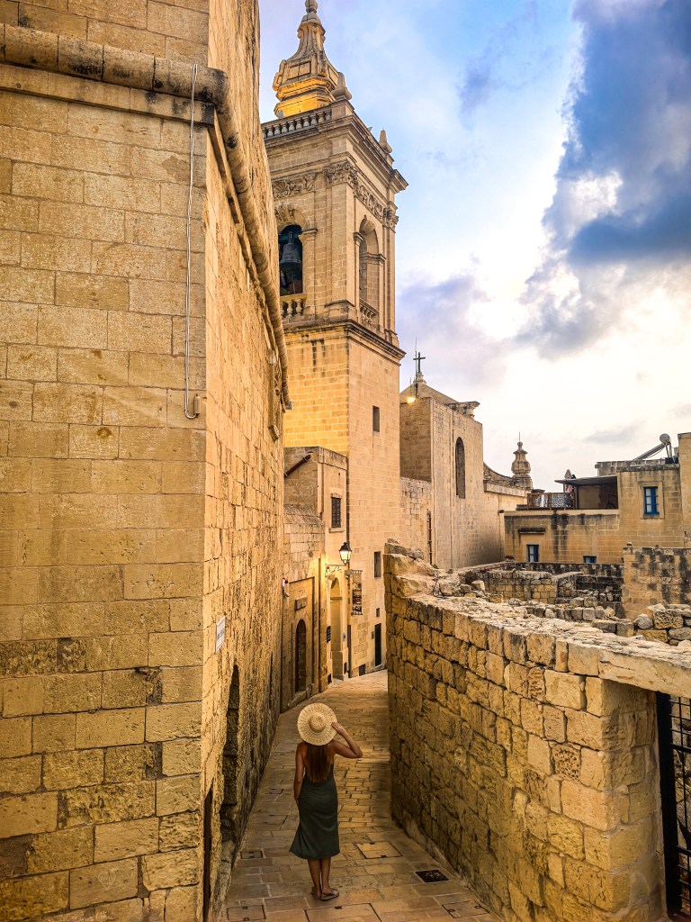 Mulher de chapéu passeia por uma rua estreita de pedra entre edifícios seculares e uma torre sineira, Cittadella, Gozo, Malta