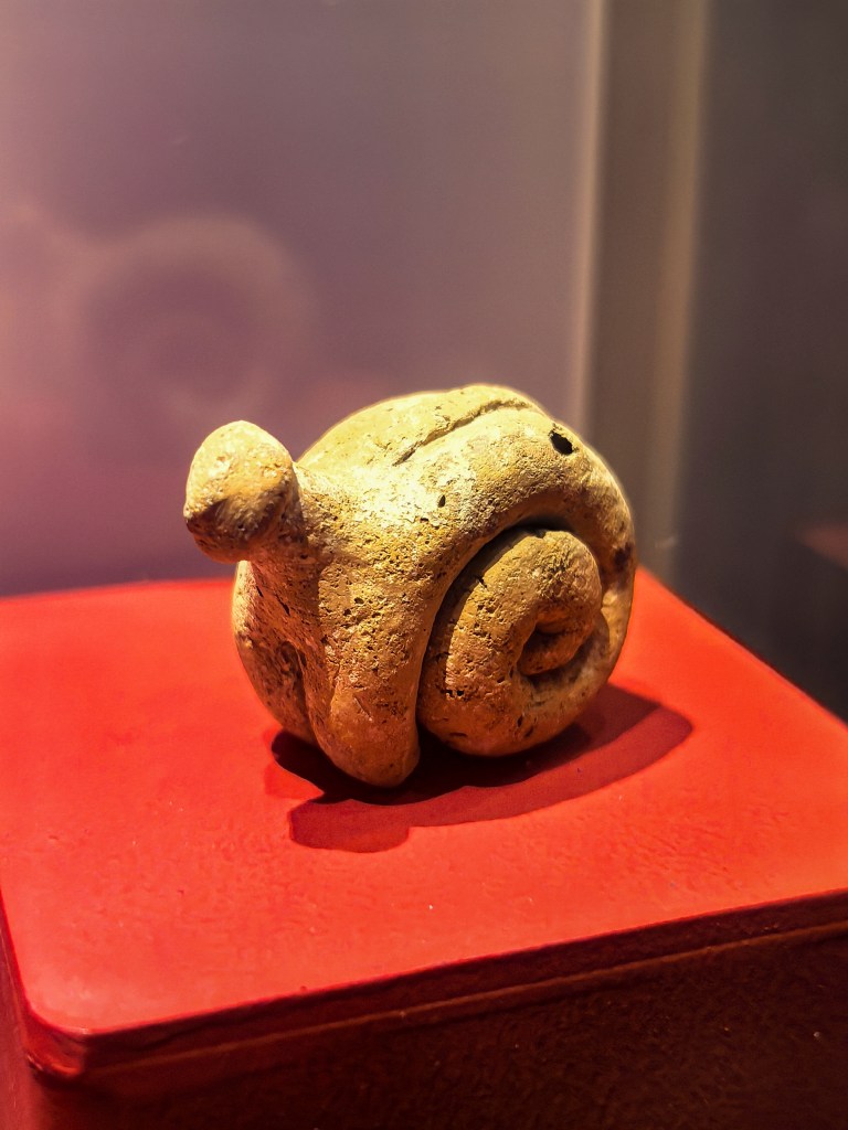 Estatueta de argila em espiral da época neolítica, encontrada nos Templos de Ġgantija, Gozo, Malta, em exposição sobre uma base vermelha