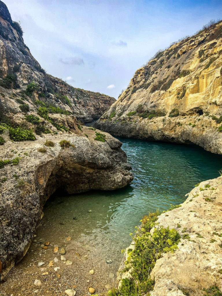 Mała kameralna plaża żwirowa w ujściu doliny il-Għasri na Gozo, otoczona stromymi wapiennymi skałami i krystalicznie czystą wodą