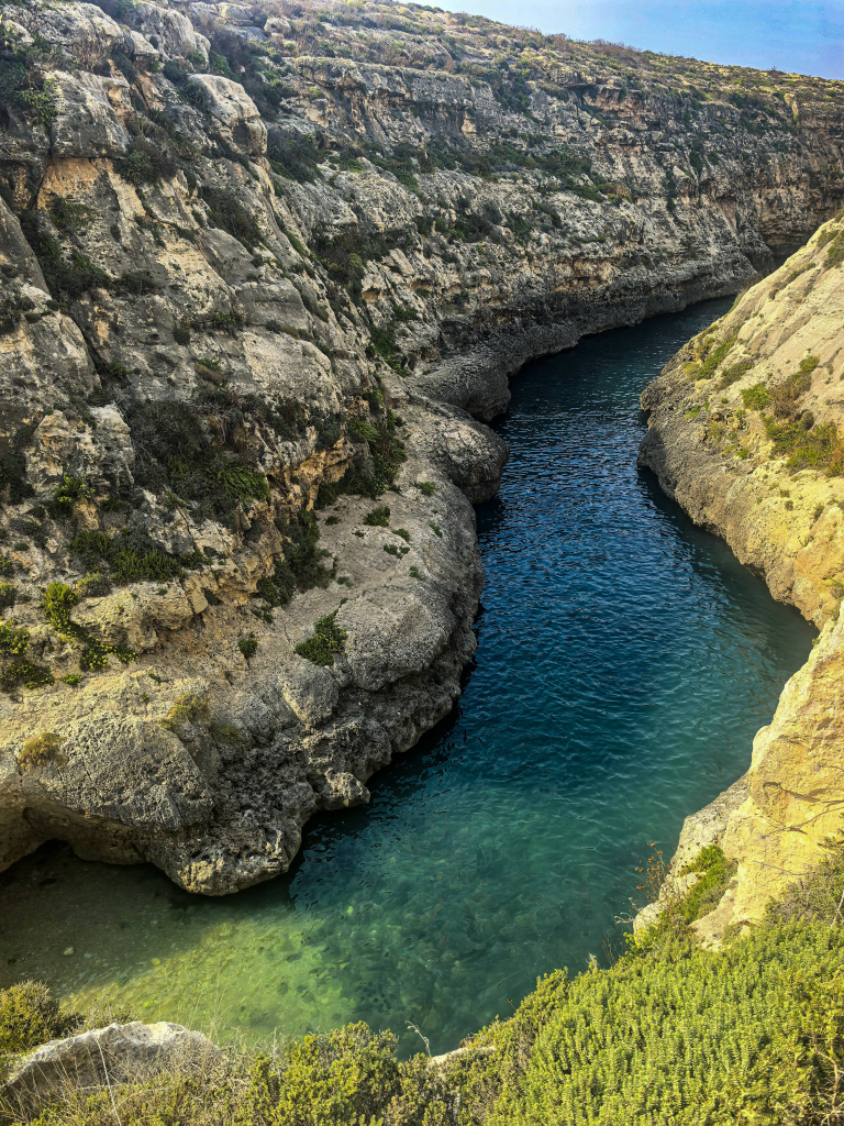Exploring Gozo in One Day – What to See Aerial view of Wied il-Ghasri canyon on Gozo, Malta, with steep limestone cliffs and turquoise water winding through the gorge.