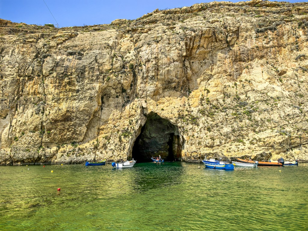 Barcos tradicionais flutuam na lagoa de águas verdes do Inland Sea com a entrada da gruta na falésia, Gozo, Malta