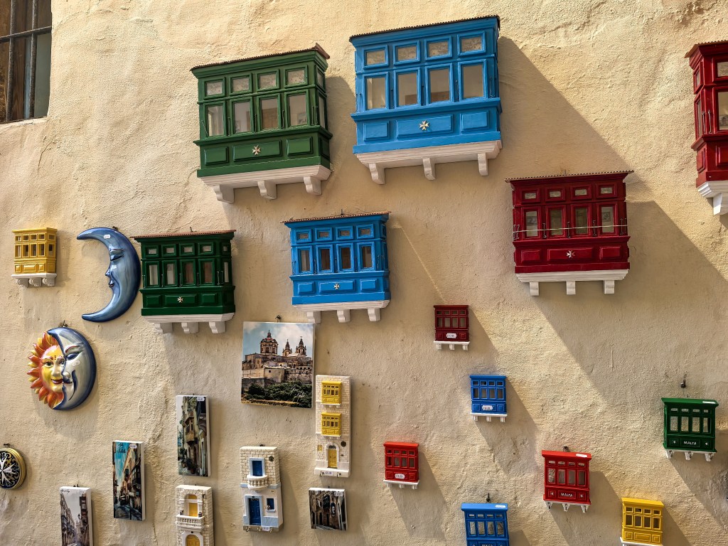 Exposição de pequenas varandas coloridas típicas de Malta, juntamente com cerâmica artesanal e fotografias, penduradas numa parede em Gozo