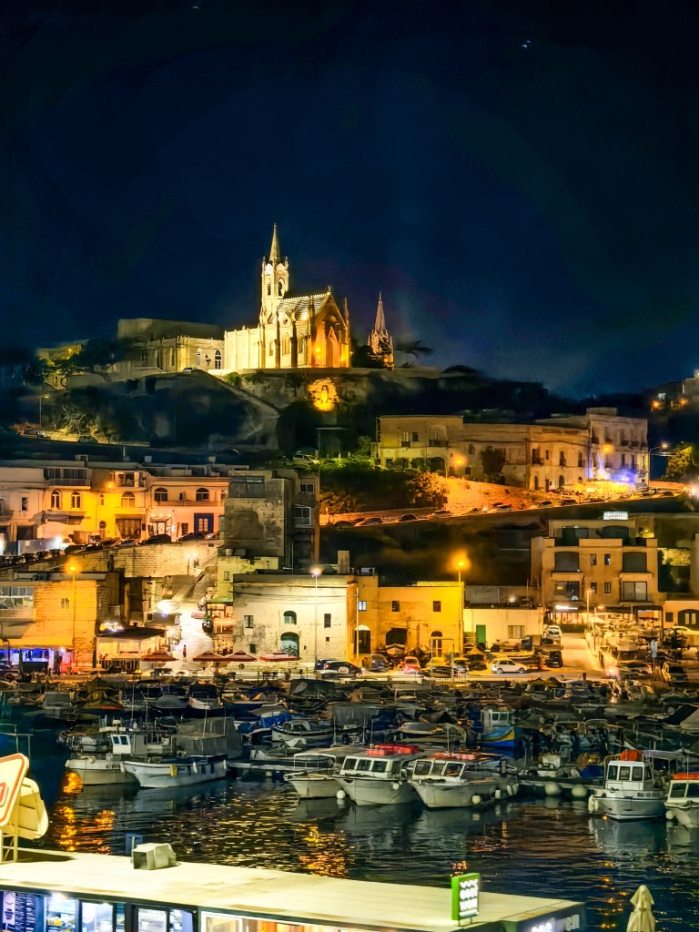 Vista noturna do porto de Mgarr com barcos atracados e, no alto, a Igreja de Nossa Senhora de Lourdes iluminada