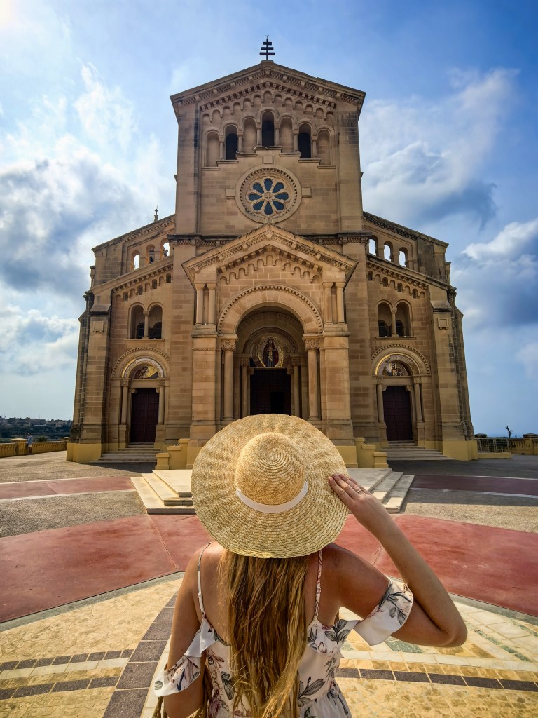 Exploring Gozo in One Day – What to See Woman in a floral dress and straw hat admiring the magnificent façade of Ta’ Pinu Basilica on Gozo, Malta – iconic pilgrimage church under a scenic sky.