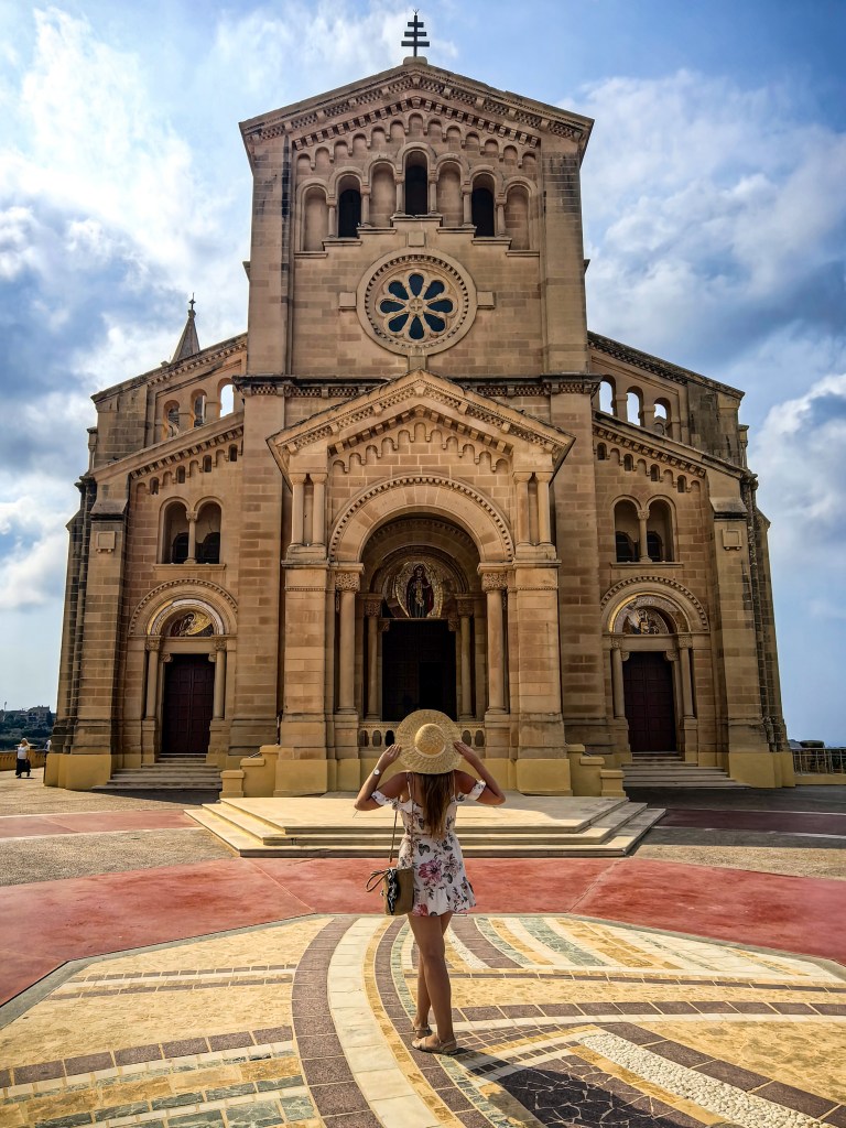 Mulher com chapéu em frente à entrada da Basílica de Ta’ Pinu, olhando para a fachada monumental