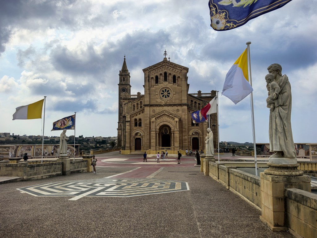 Exploring Gozo in One Day – What to See Panoramic view of Ta' Pinu Basilica courtyard on Gozo, Malta, with waving flags, white statues, and pilgrims approaching the iconic church under a dramatic sky.