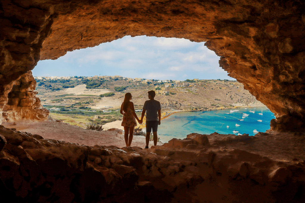 Casal na entrada da gruta Tal-Mixta, com vista panorâmica para a praia de areia vermelha e mar azul da Baía de Ramla, em Gozo