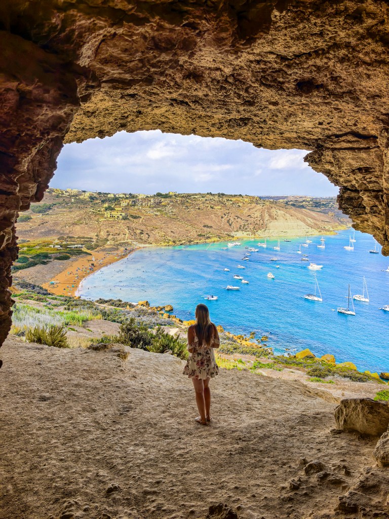 Mulher de vestido de verão na entrada da gruta Tal-Mixta, olhando para a baía de Ramla com areia vermelha e mar azul-turquesa em Gozo, Malta