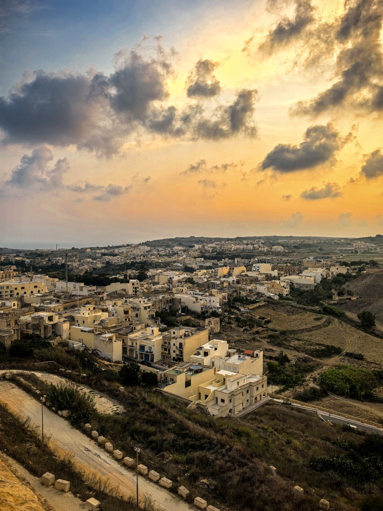 Paisagem urbana de Victoria, com casas e campos ao pôr do sol, vista desde as muralhas da Cittadella