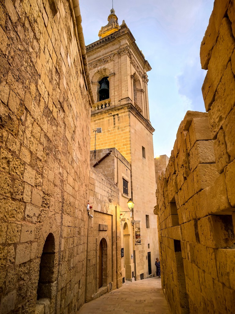 Rua estreita de Victoria ladeada por muralhas históricas em pedra e vista para a torre sineira da catedral