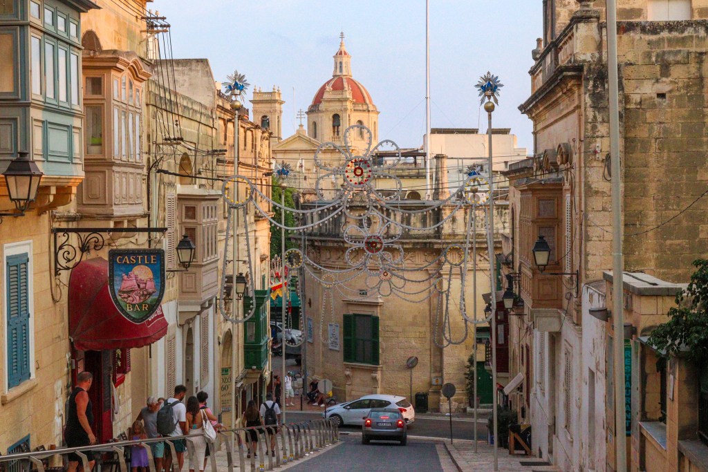 Rua antiga de Victoria (Rabat) decorada para festas, com varandas típicas maltesas e vista para cúpulas de igrejas