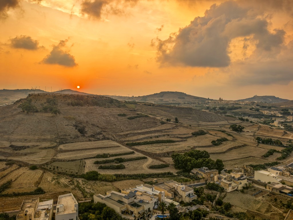 Exploring Gozo in One Day – What to See Panoramic sunset over terraced fields and hills near Victoria, Gozo, Malta, with orange clouds and rural buildings in the foreground