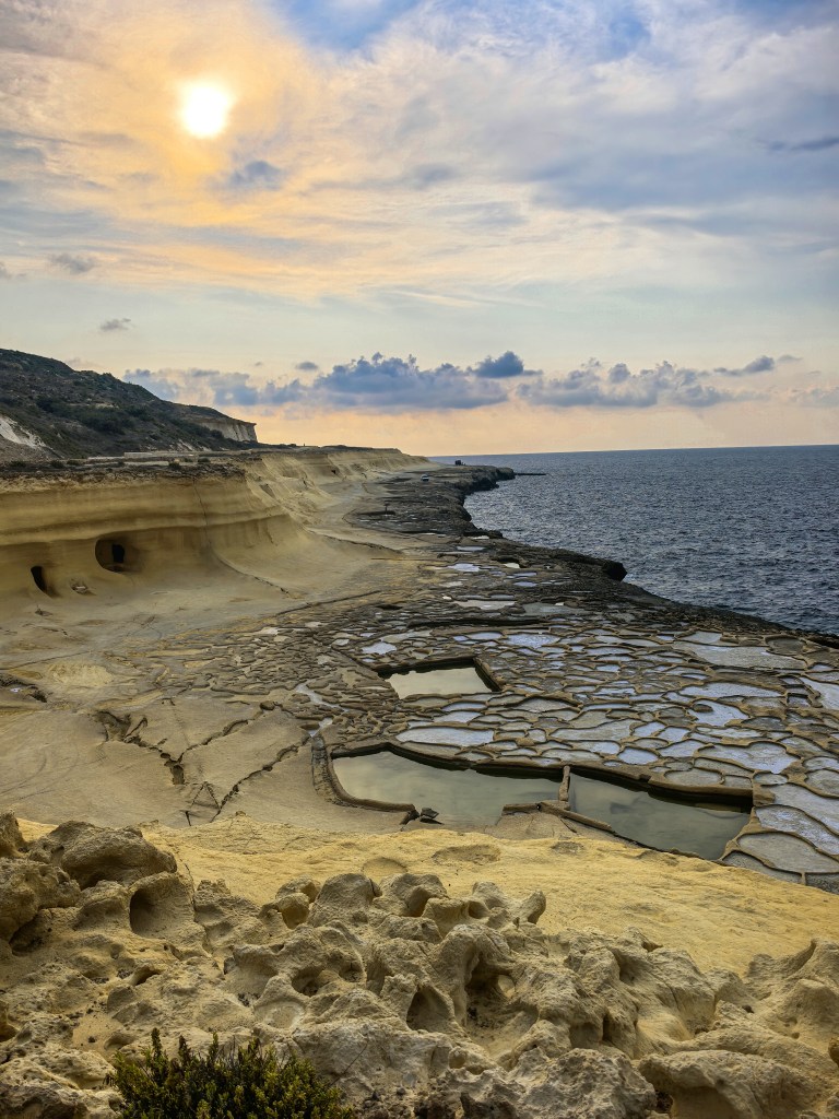 Salinas tradicionais de Xwejni iluminadas pelo sol poente, com céu colorido e mar ao fundo, Gozo, Malta
