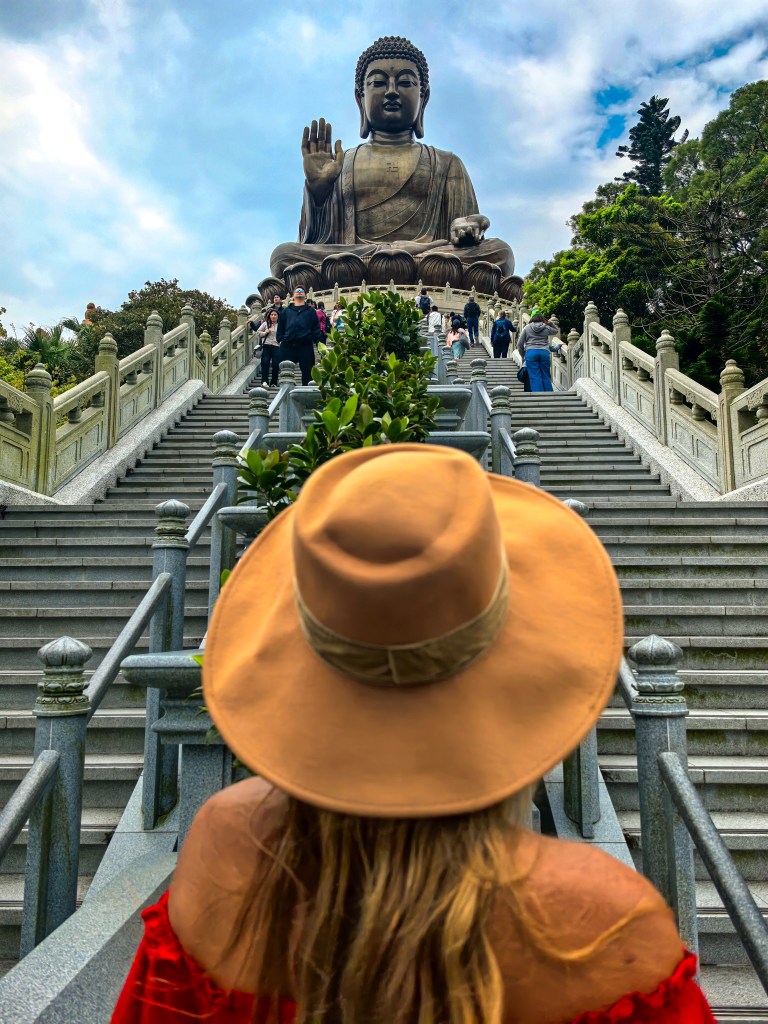 Mulher de chapéu e vestido vermelho de costas para a escadaria com o Grande Buda Tian Tan ao fundo, cercada por plantas, Hong Kong.