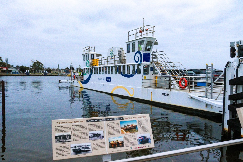Raymond Island – O Guia Completo da Ilha dos Coalas 🐨 Ferry de carros e passageiros no terminal de Raymond Island, Austrália, com painel informativo à frente.