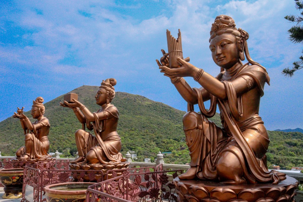 Bronze Bodhisattva statues holding offerings with mountains in the background, near Tian Tan Buddha, Hong Kong.