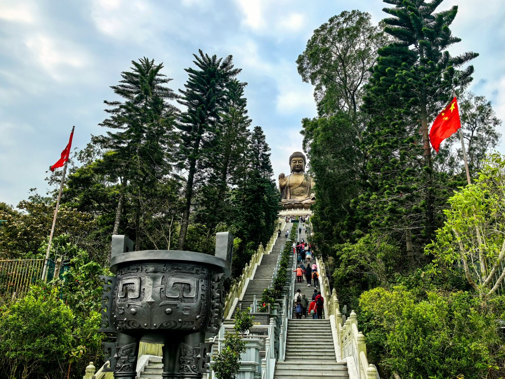 Long staircase leading to Tian Tan Buddha statue surrounded by trees and Chinese flags, Hong Kong.