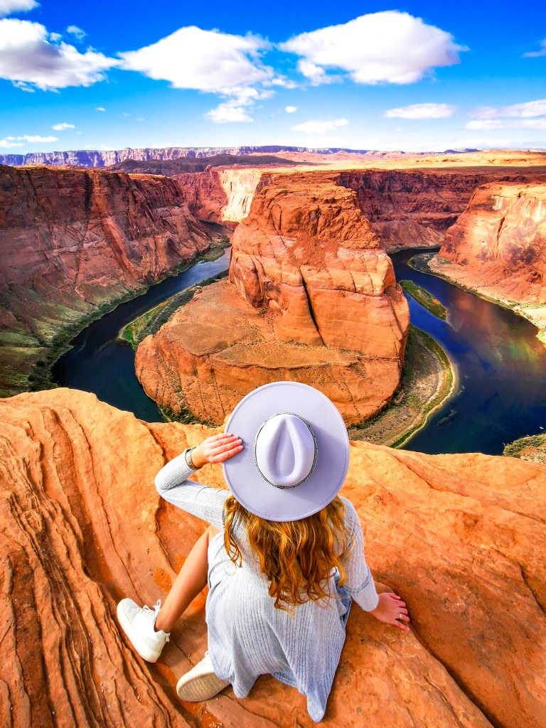 Kobieta w jasnej sukience i kapeluszu siedzi na skale nad przepaścią, patrząc na zakole rzeki Kolorado w Horseshoe Bend.