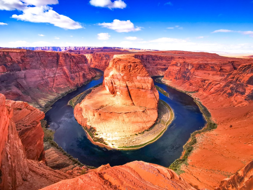 Intensywnie pomarańczowe skały Horseshoe Bend otaczające zakole rzeki Kolorado, błękitne niebo z chmurami nad kanionem.