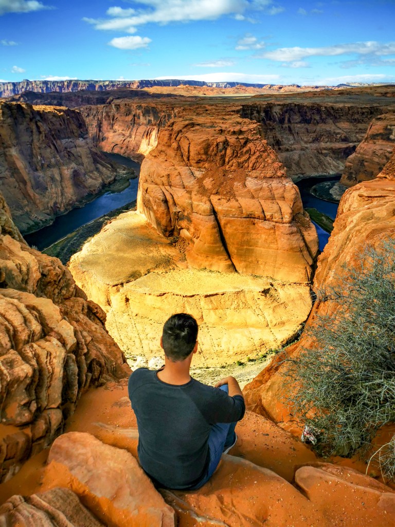 Widok na Horseshoe Bend w Arizonie, z osobą siedzącą na krawędzi kanionu, otoczony czerwonymi skałami i meandrującą rzeką Kolorado w dole.