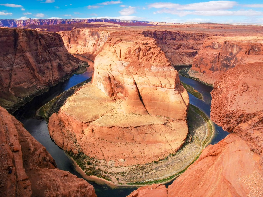 Widok na Horseshoe Bend, naturalne zakole rzeki Kolorado otoczone czerwonymi skałami i zielonymi roślinami, na tle błękitnego nieba.