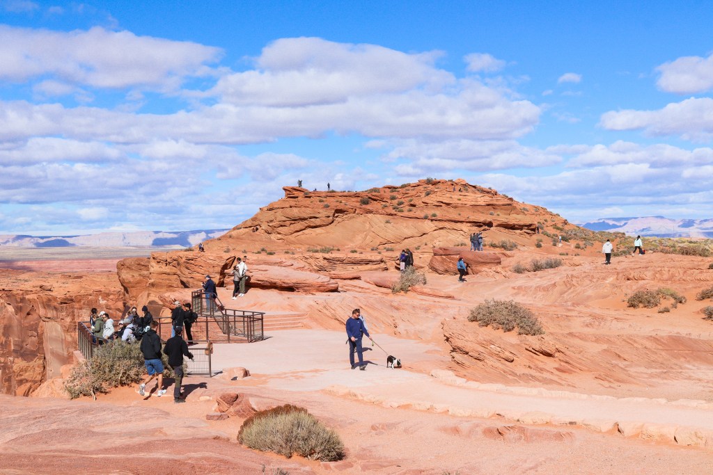 Widok na punkt widokowy Horseshoe Bend, z turystami podziwiającymi majestatyczny kanion oraz pomarańczowe formacje skalne w Arizonie, z błękitnym niebem w tle.