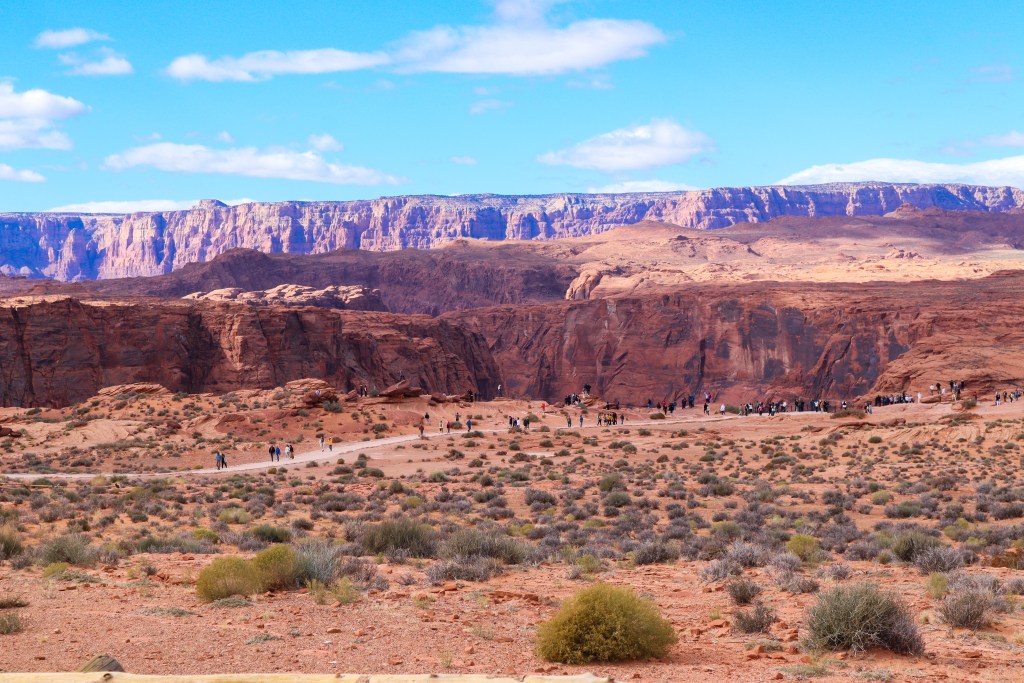 Widok na pustynny teren i ścieżkę pełną turystów prowadzącą do punktu widokowego Horseshoe Bend, na tle monumentalnych ścian kanionu. W tle widać majestatyczne kaniony oraz błękitne niebo.