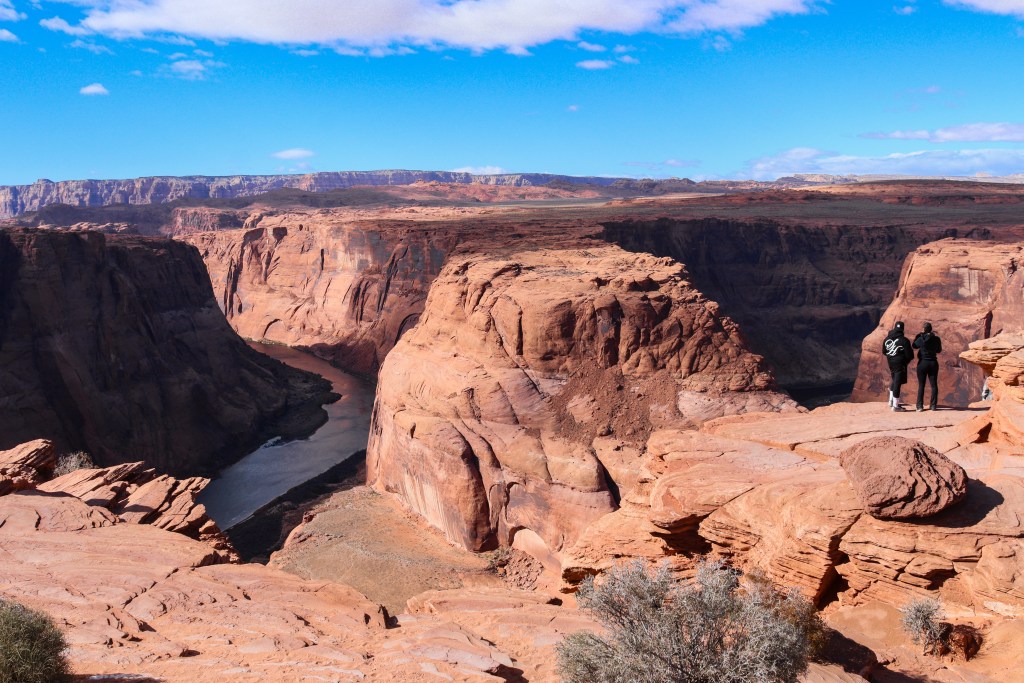 Widok na Horseshoe Bend w Arizonie, z rzeką Kolorado wijącą się przez czerwone skały kanionu. Dwie osoby stoją na krawędzi, podziwiając spektakularny krajobraz.