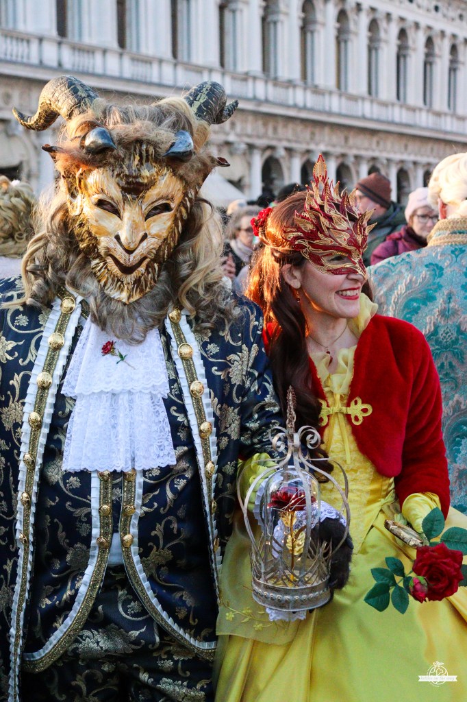 Casal do Carnaval de Veneza como Bela e Monstro: besta cornuda em fato azul e Bela de amarelo com rosa em redoma de vidro