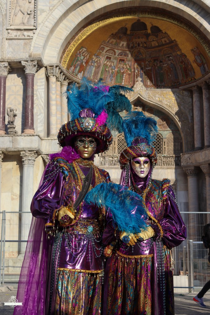Casal em fatos orientais roxos brilhantes do Carnaval de Veneza com penas junto aos arcos da Basílica de São Marcos