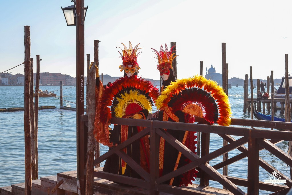 Casal do Carnaval de Veneza em fatos laranja e preto com penas e leques grandes junto ao cais de madeira sobre a lagoa