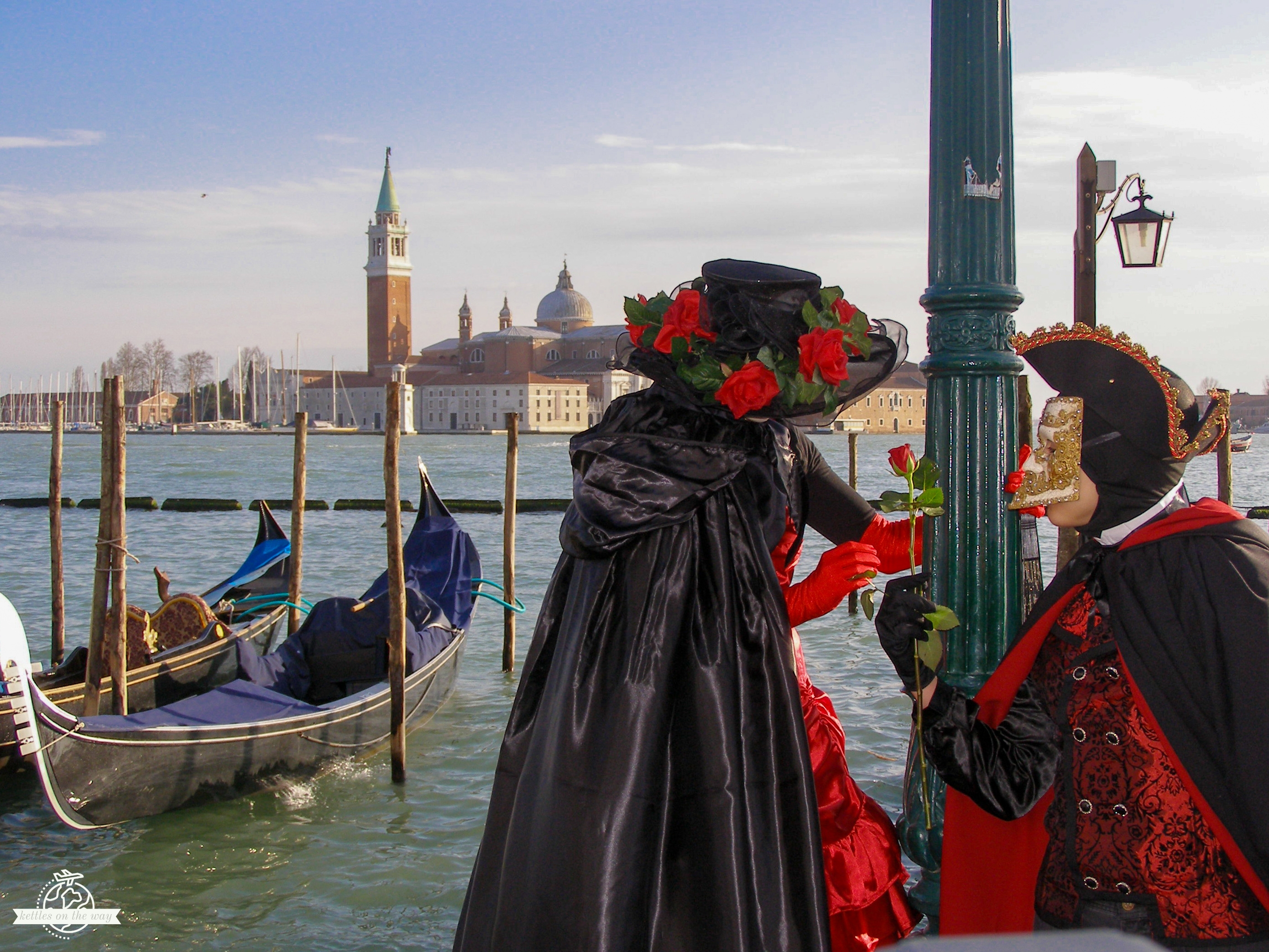 Veneza: Tudo o que deves saber antes da tua viagem Casal romântico do Carnaval de Veneza em fatos vintage e máscaras junto ao Canal Grande com gondolas e igreja ao pôr do sol