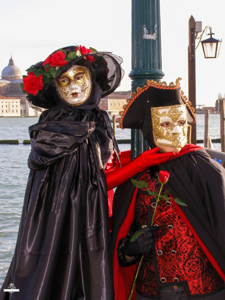 Casal romântico do Carnaval de Veneza em capa preta e fato vermelho a trocar uma rosa junto à lagoa com cúpula de igreja