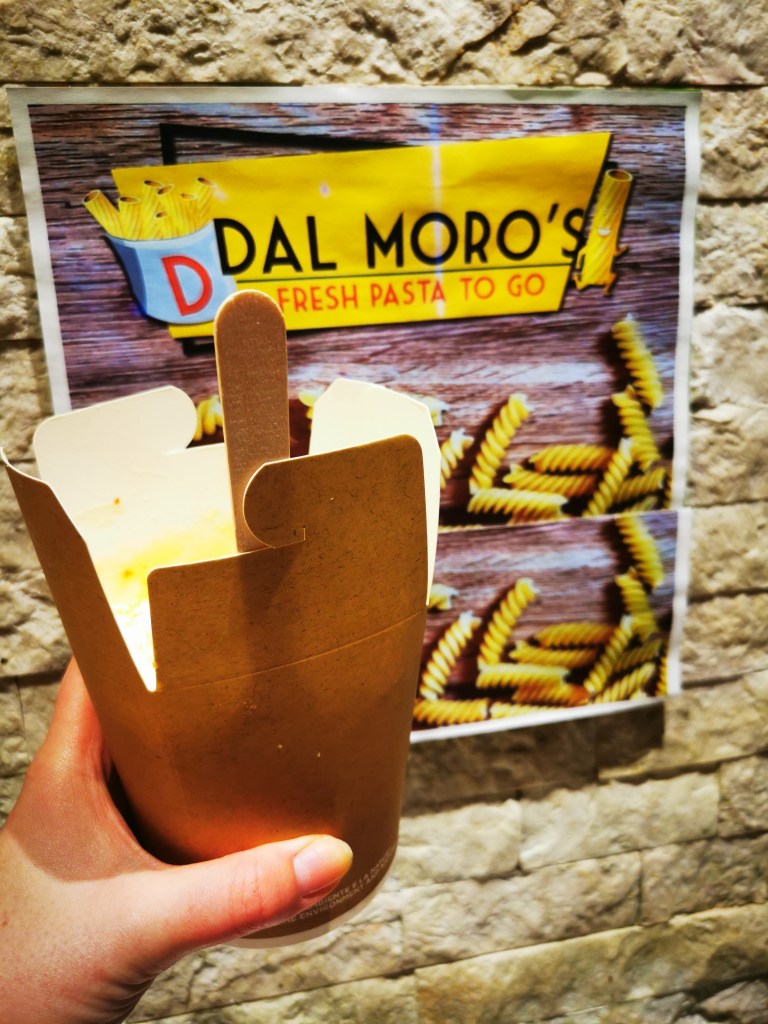 Hand holding takeaway box of Dal Moro's fresh pasta with spaghetti against colorful shop sign on stone wall
