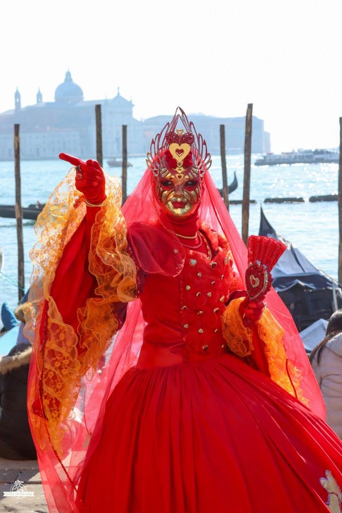 Woman in bright red Venice Carnival dress with lace sleeves and heart accessories posing by the lagoon and gondolas