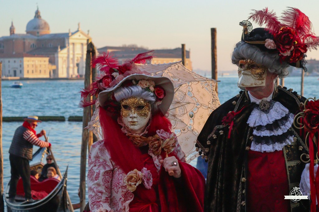 Máscaras elegantes do Carnaval de Veneza em fatos pretos e vermelhos com rosas perto de gôndolas e igreja de São Jorge Maggiore