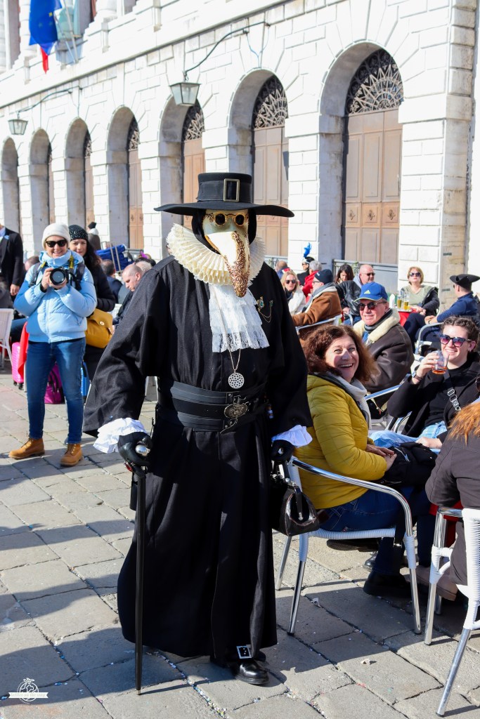Médico da peste icónico com chapéu de aba larga e máscara de bico numa rua do Carnaval de Veneza perto de edifício branco histórico