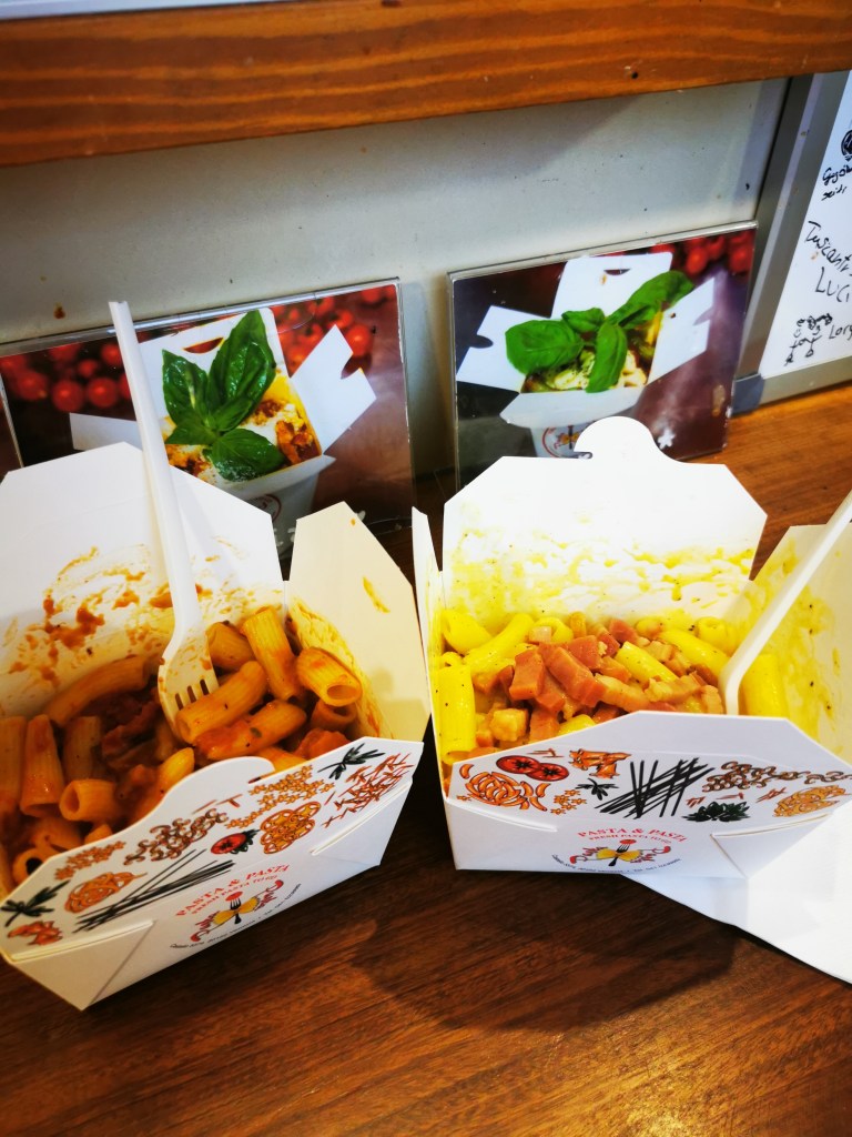 Duas caixas de takeaway do Dal Moro: penne cremoso amarelo e rigatoni de tomate numa mesa de madeira com manjericão