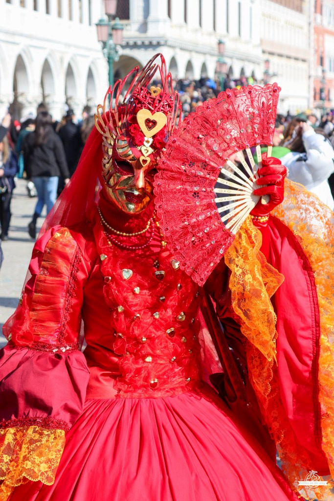 Vibrant red gown lady with heart-adorned mask and lace fan posing in Venice Carnival crowd by arched buildings