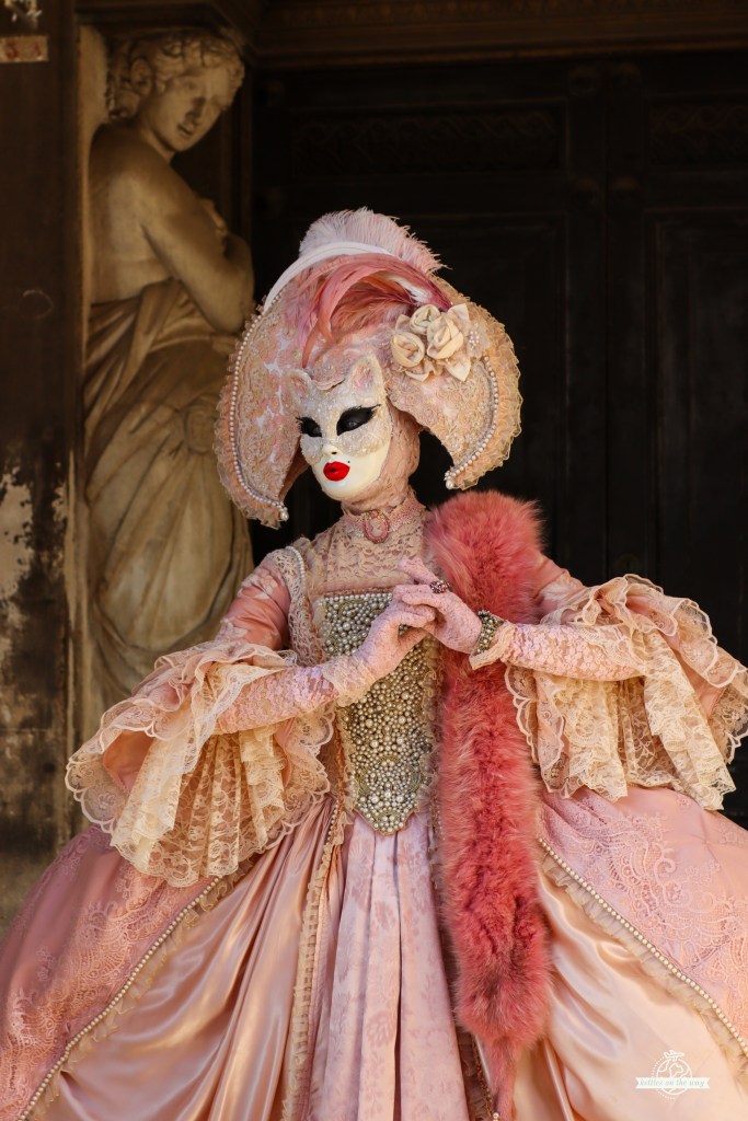 Senhora elegante em vestido rosa pastel ornamentado do Carnaval de Veneza com chapéu largo com penas junto a porta histórica