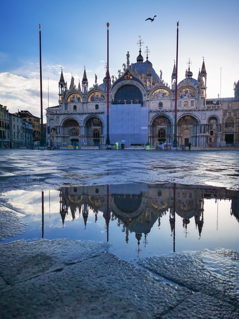 Widok na Bazylikę św. Marka w Wenecji odbijającą się w wodzie po deszczu, z pięknymi detalami architektonicznymi i niebem w tle.