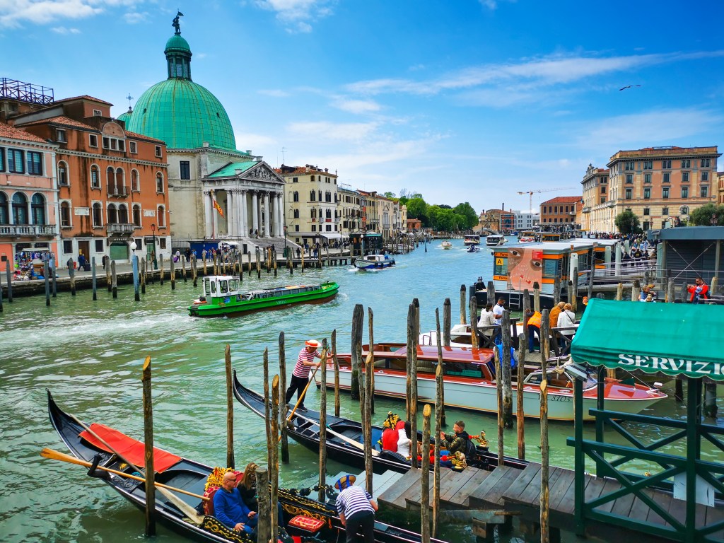 Gondolierzy i vaporetto na Canal Grande w Wenecji przy przystani, z zielonymi wodami kanału i charakterystyczną zieloną kopułą kościoła San Simeon Piccolo.