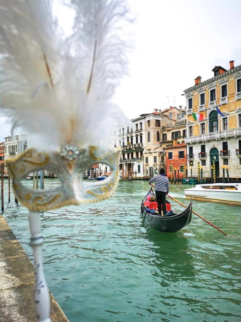 Widok na weneckiego gondoliera płynącego po Canal Grande, z białą maską w stylu karnawałowym z piórami na pierwszym planie.