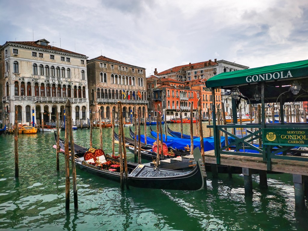 Widok na przystań z gondolami w Wenecji przy Canal Grande, otoczoną kolorowymi budynkami i wodą.
