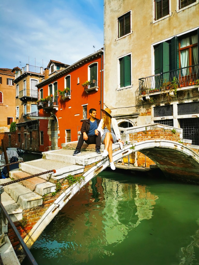 Para na moście bez barierek Ponte Chiodo w dzielnicy Cannaregio w Wenecji, z zielonymi wodami kanału i weneckimi budynkami w otoczeniu.