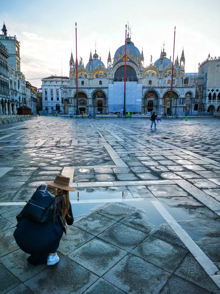 Kobieta w kapeluszu klęczy na placu, fotografując odbicie w kałuży, z widokiem na bazylikę Świętego Marka w Wenecji.